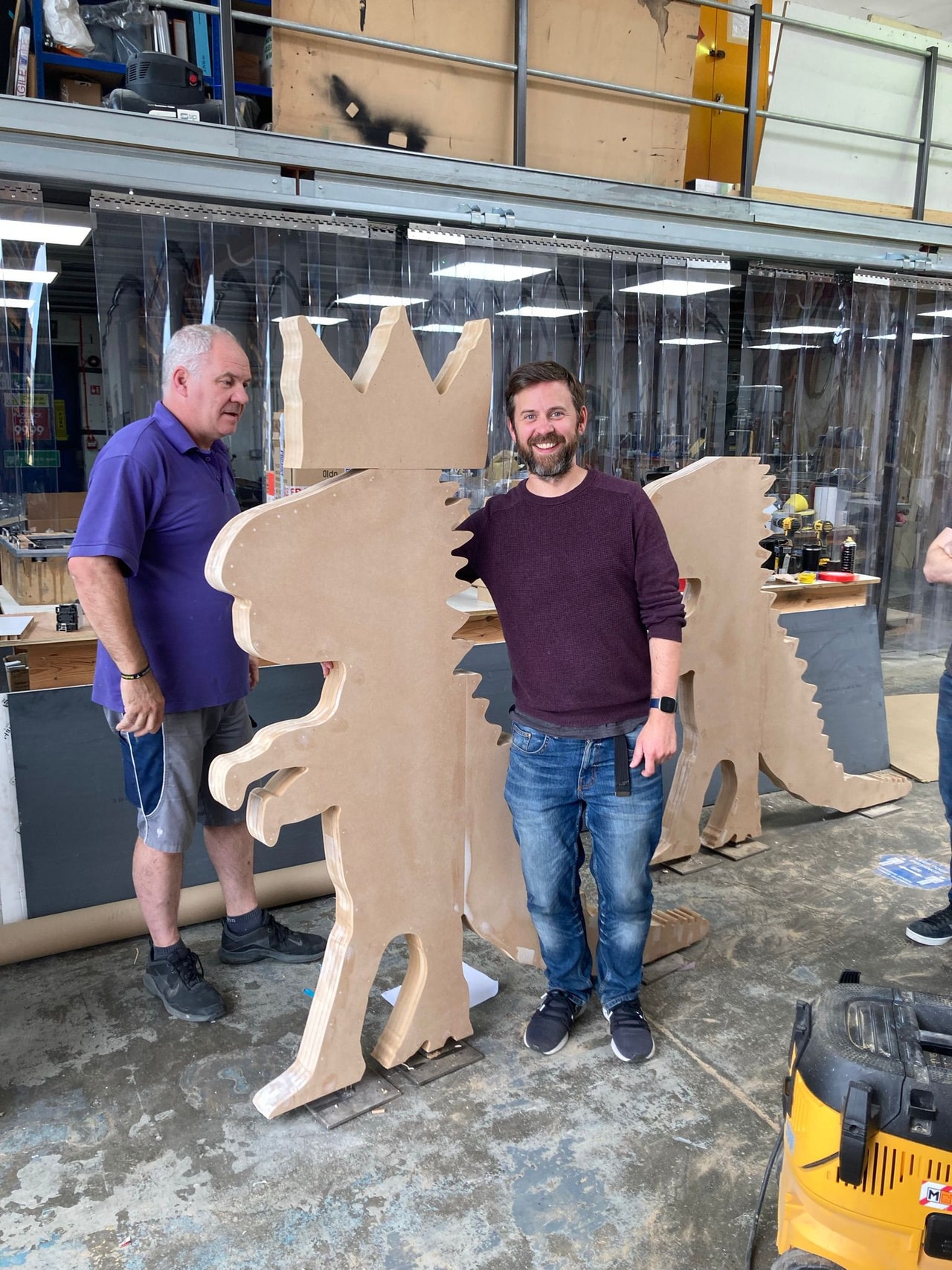 Workshop photo showing large CNC-cut MDF character cutouts in a fabrication workshop with staff and tools in the background.