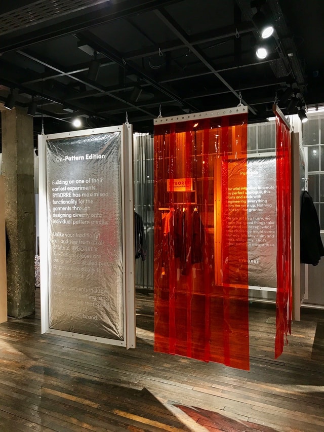 Indoor exhibition/pop-up retail installation with printed display panels and red translucent strip curtains showcasing garments in a gallery-style space.