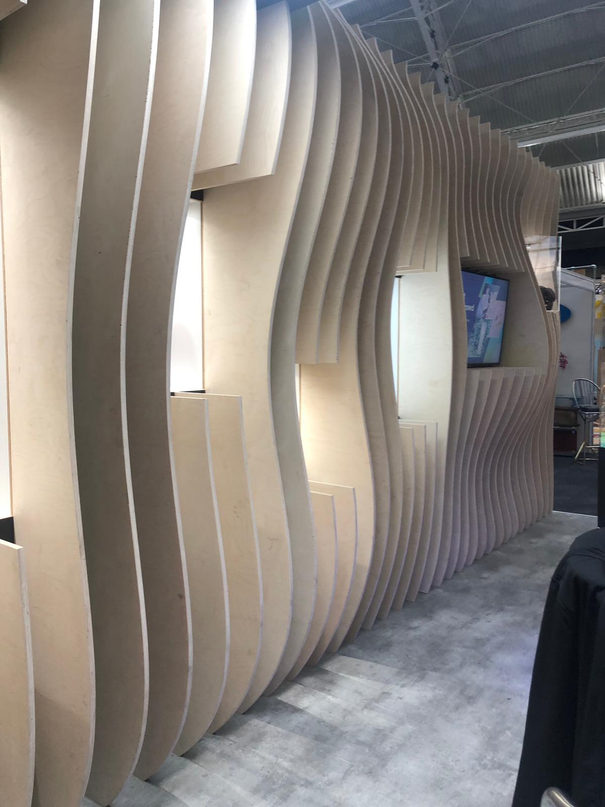 Curved, layered plywood exhibition wall composed of CNC-cut ribs with an integrated flat-screen, forming a sculptural trade show booth partition.