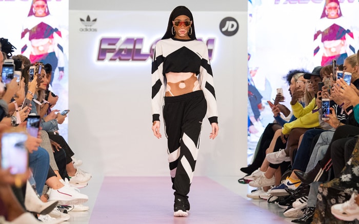 Runway fashion show with a model walking down the catwalk, seated audience on both sides and a branded printed backdrop behind the stage.