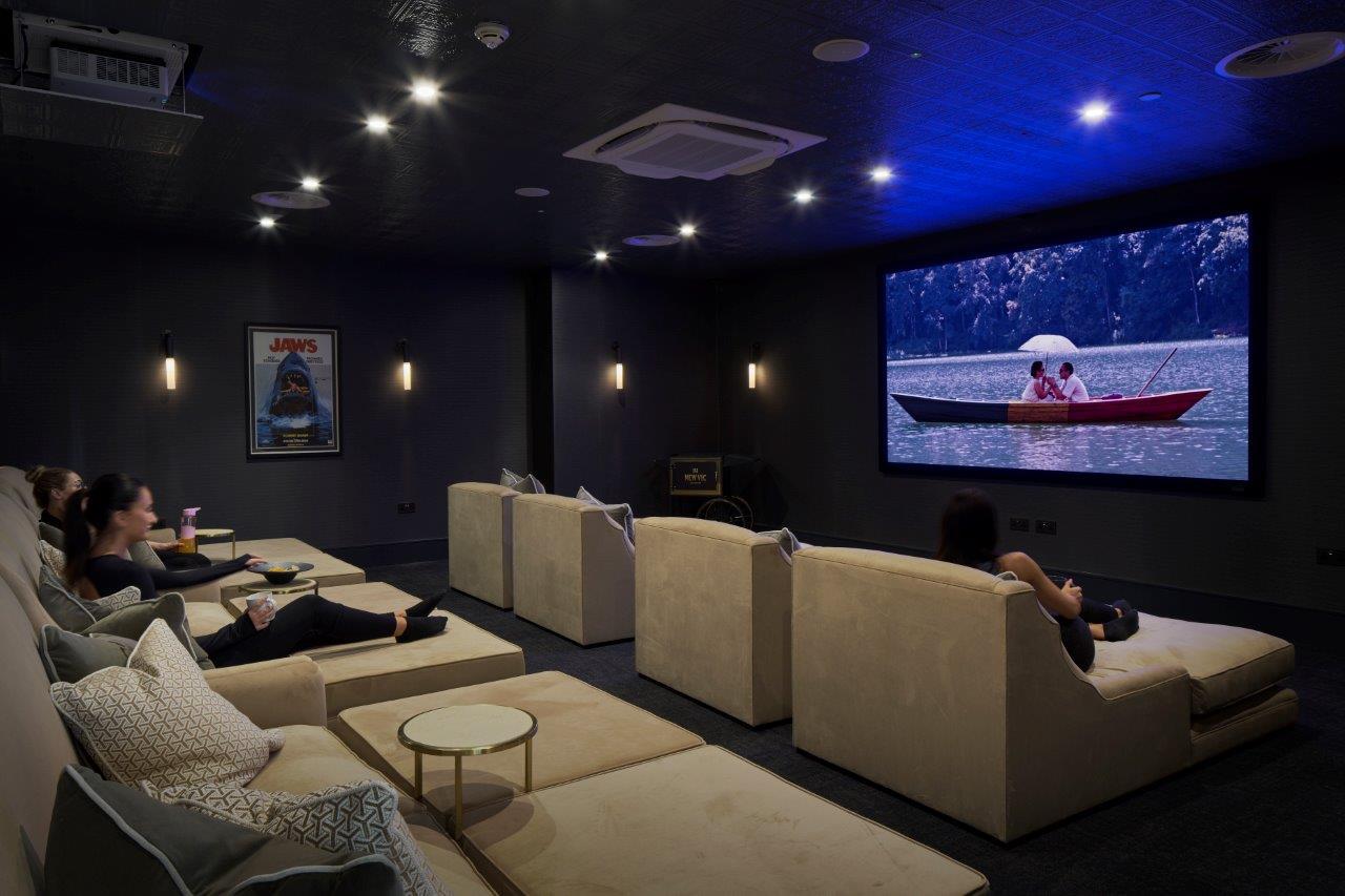 Interior screening/event space with plush tiered seating and a large projection screen, dimmable downlights and framed film poster for a premium cinema experience.
