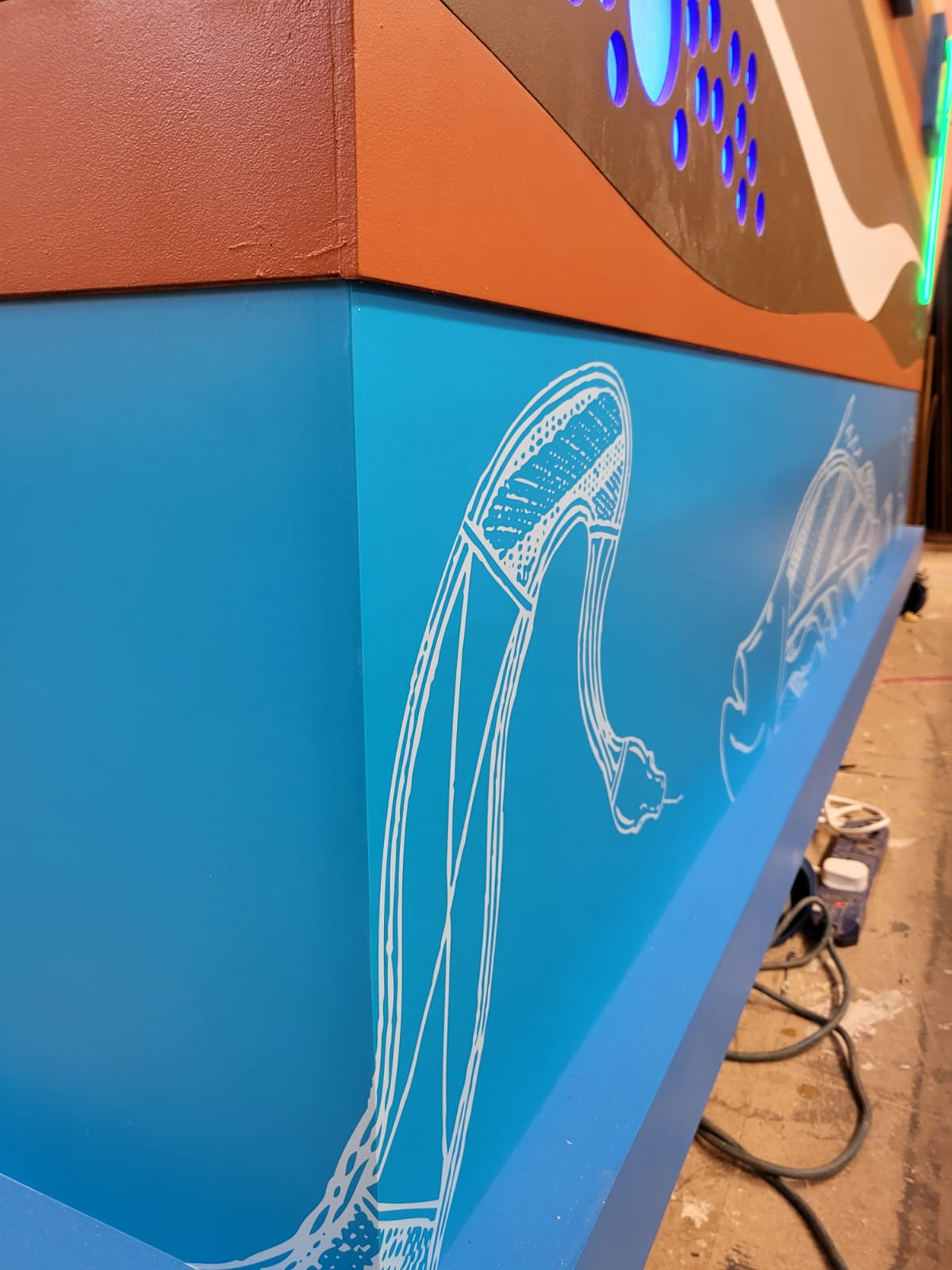 Workshop shot of a blue painted display panel with white illustrative graphics and integrated LED/neon lighting, photographed during finishing/installation work.