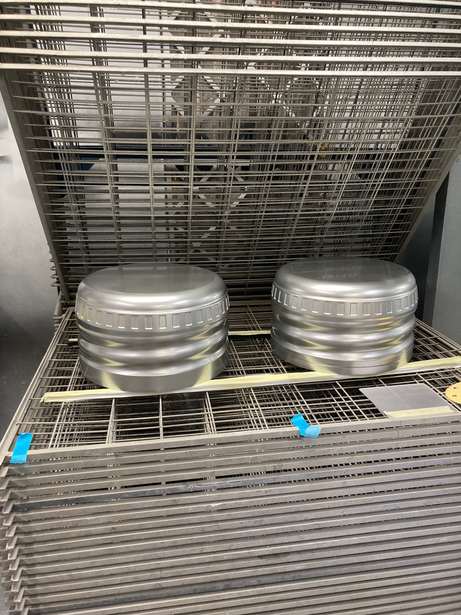 Two silver-painted metal caps drying on a wire rack inside a finishing booth, showing a paint/coating process.