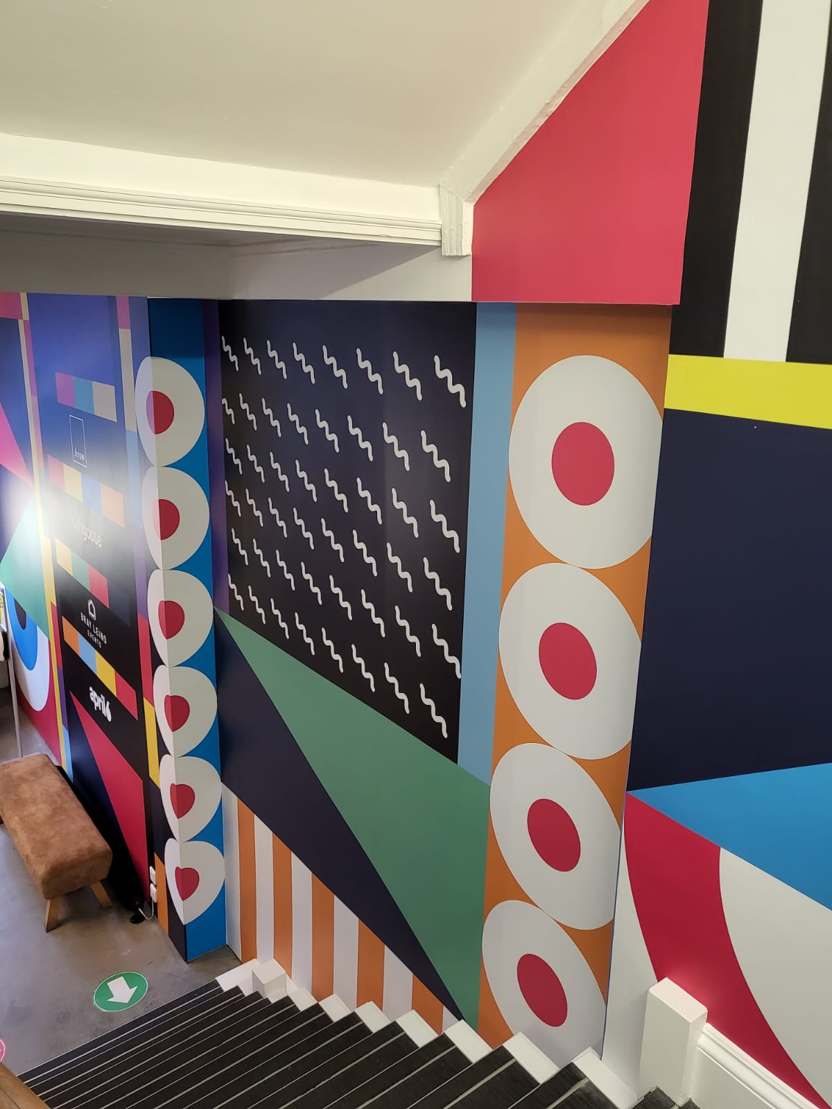 Bright geometric large-format printed wall graphics installed on an interior stairwell in a retail/office environment, featuring bold shapes and colours.