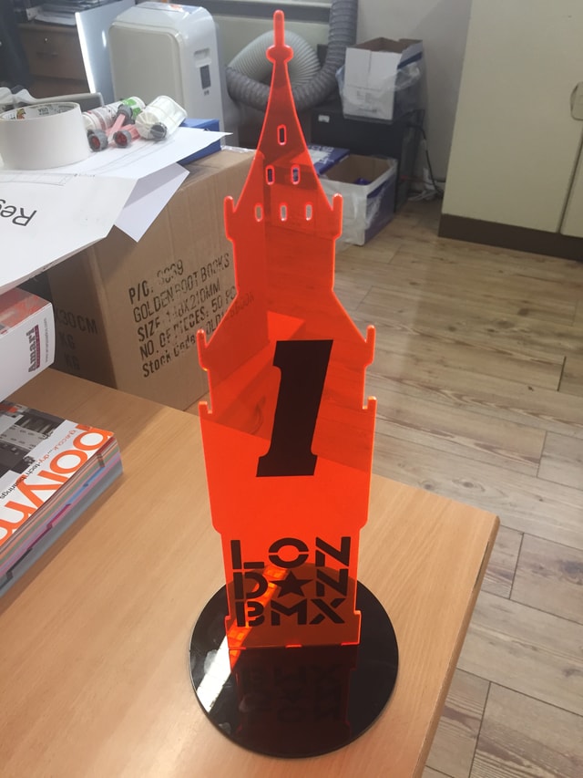 Orange laser-cut acrylic London BMX trophy on a black base photographed on a workshop bench; finished display piece with cut lettering and decorative tower shape.