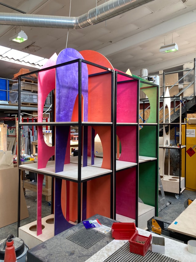 Colourful modular display shelving units being assembled in a fabrication workshop, showing painted plywood panels and metal framing for retail fixtures.