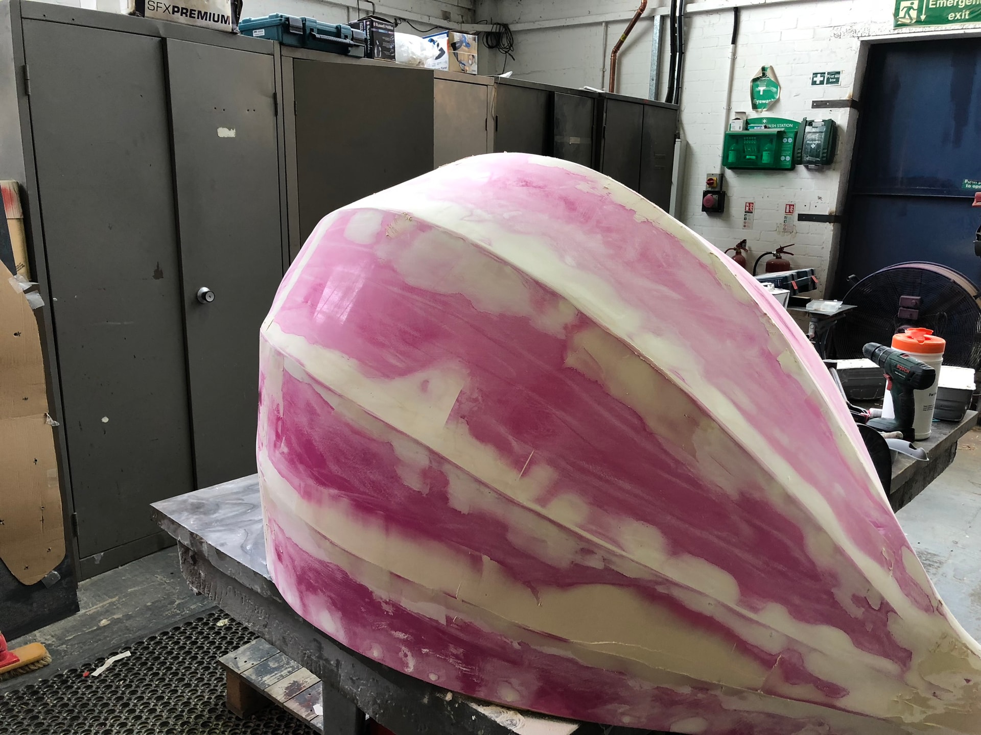 Workshop photo of a large formed plastic shell undergoing filler and sanding on a workbench, with tools and cabinets visible in the background.
