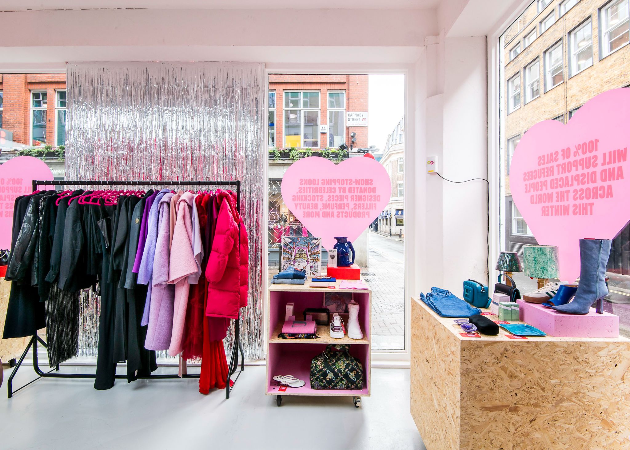 Bright retail interior / pop-up shop showing clothing rail, product displays on OSB plinths and shelving, with pink heart window vinyl graphics and a silver fringe backdrop.