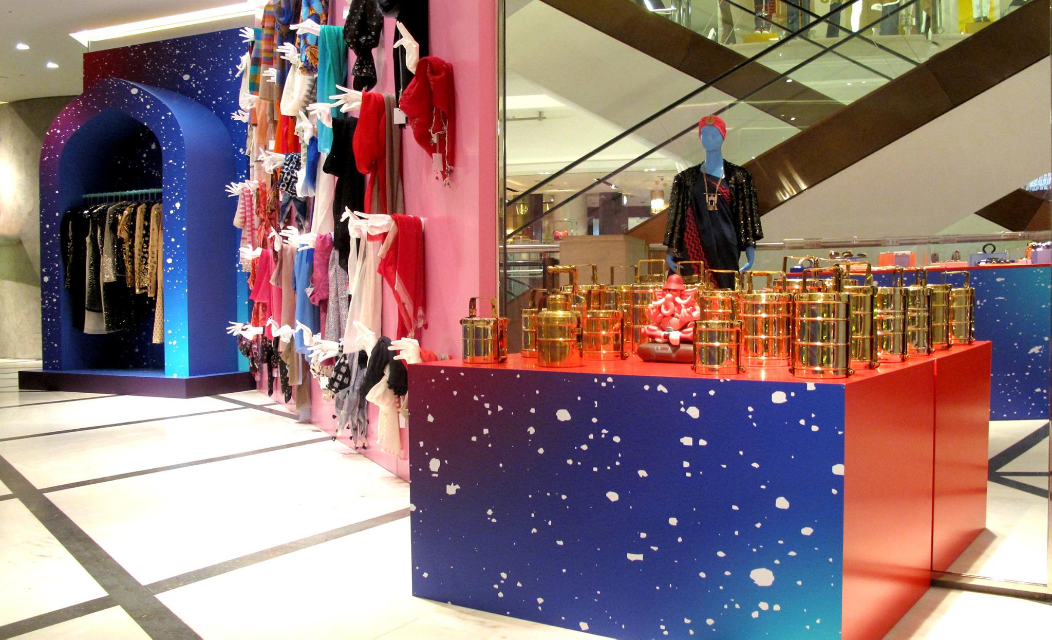 Colorful in-store retail display featuring mannequins, wall-mounted garments and speckled blue display islands with product tins inside a department store setting.