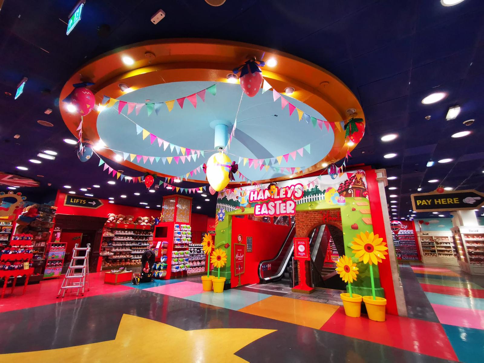 Colorful Hamleys store interior with a seasonal 'Hamleys Easter' arch over an escalator, product displays and clear directional signage in a bright retail environment.