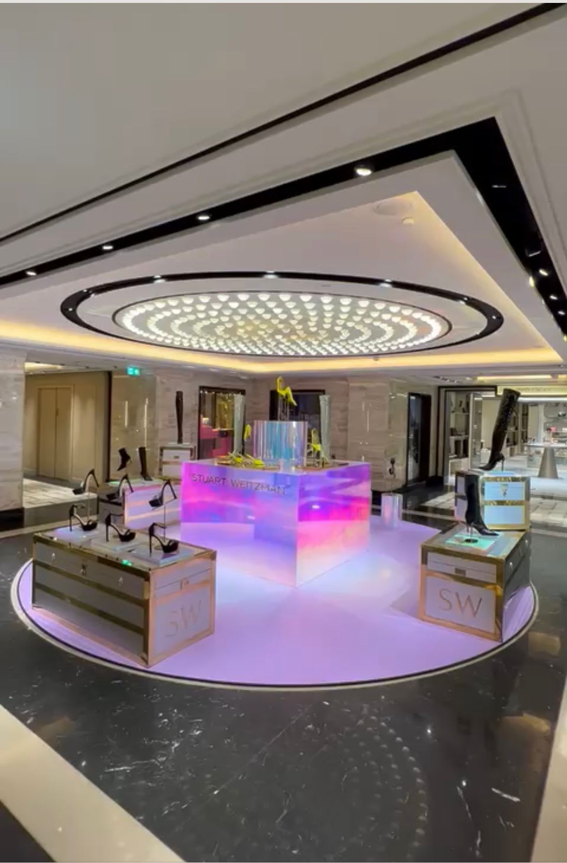 Iridescent illuminated retail display island with Stuart Weitzman branding and product pedestals in a premium department store setting.