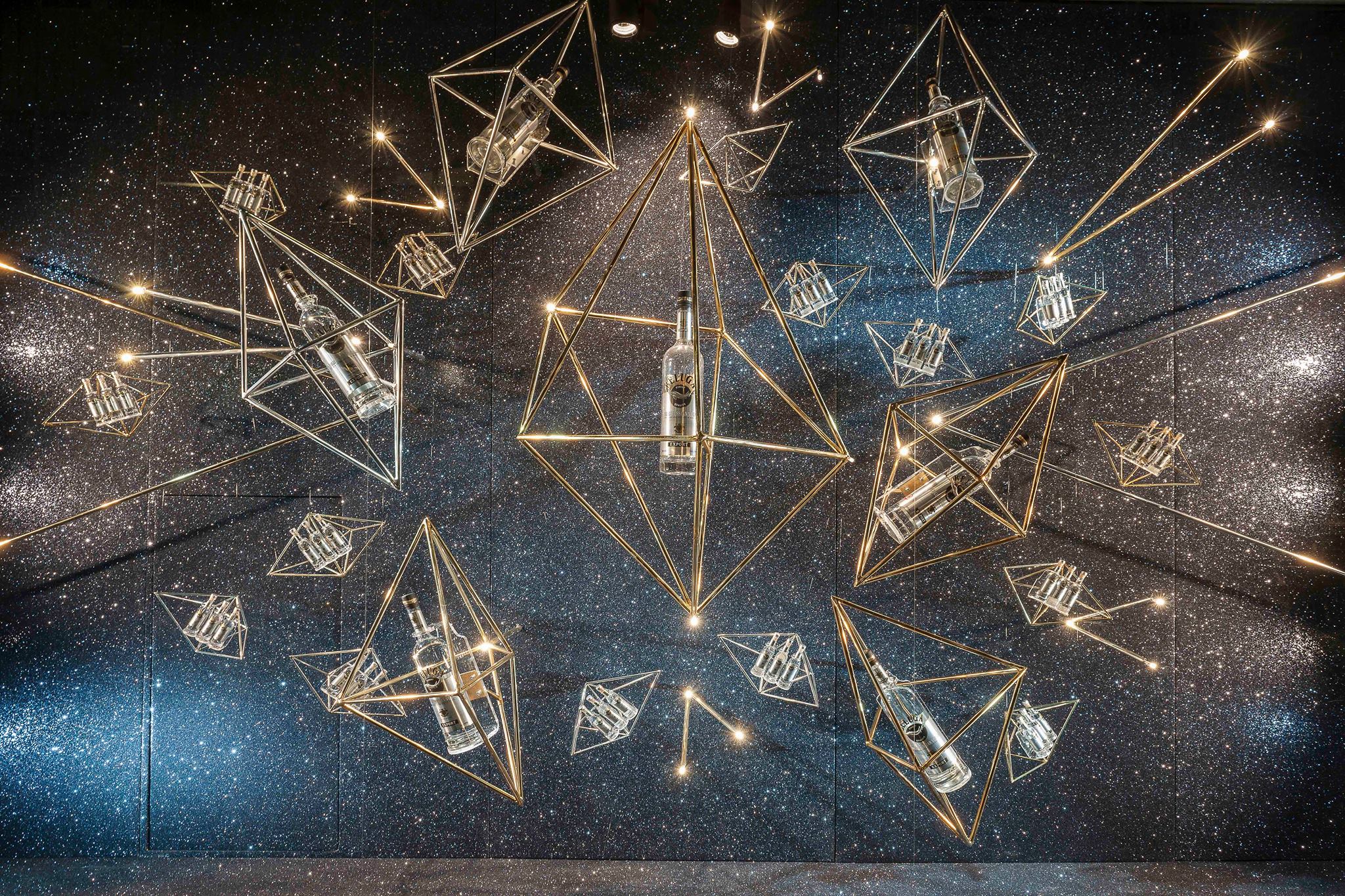 Ceiling-mounted geometric metal display holding glass bottles against a sparkling starfield backdrop, an editorial retail fixture or window installation that creates an immersive product display.