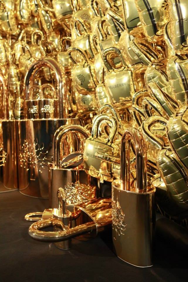 Luxury Mulberry retail/window display featuring an installation of gold padlocks sculptures and reflective metallic balloons, styled as a high-end merchandising feature.