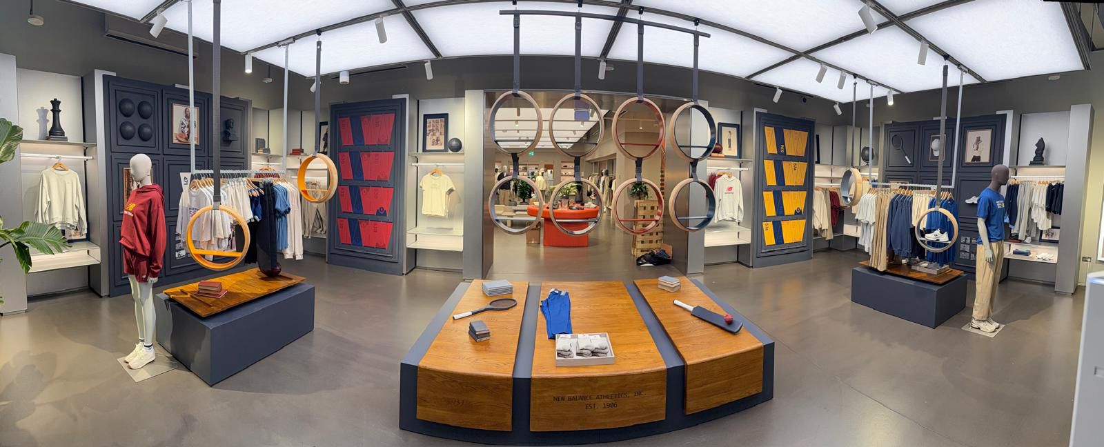 Bright, professionally fitted New Balance retail interior with mannequins, hanging ring displays and timber display tables showcasing apparel and footwear.