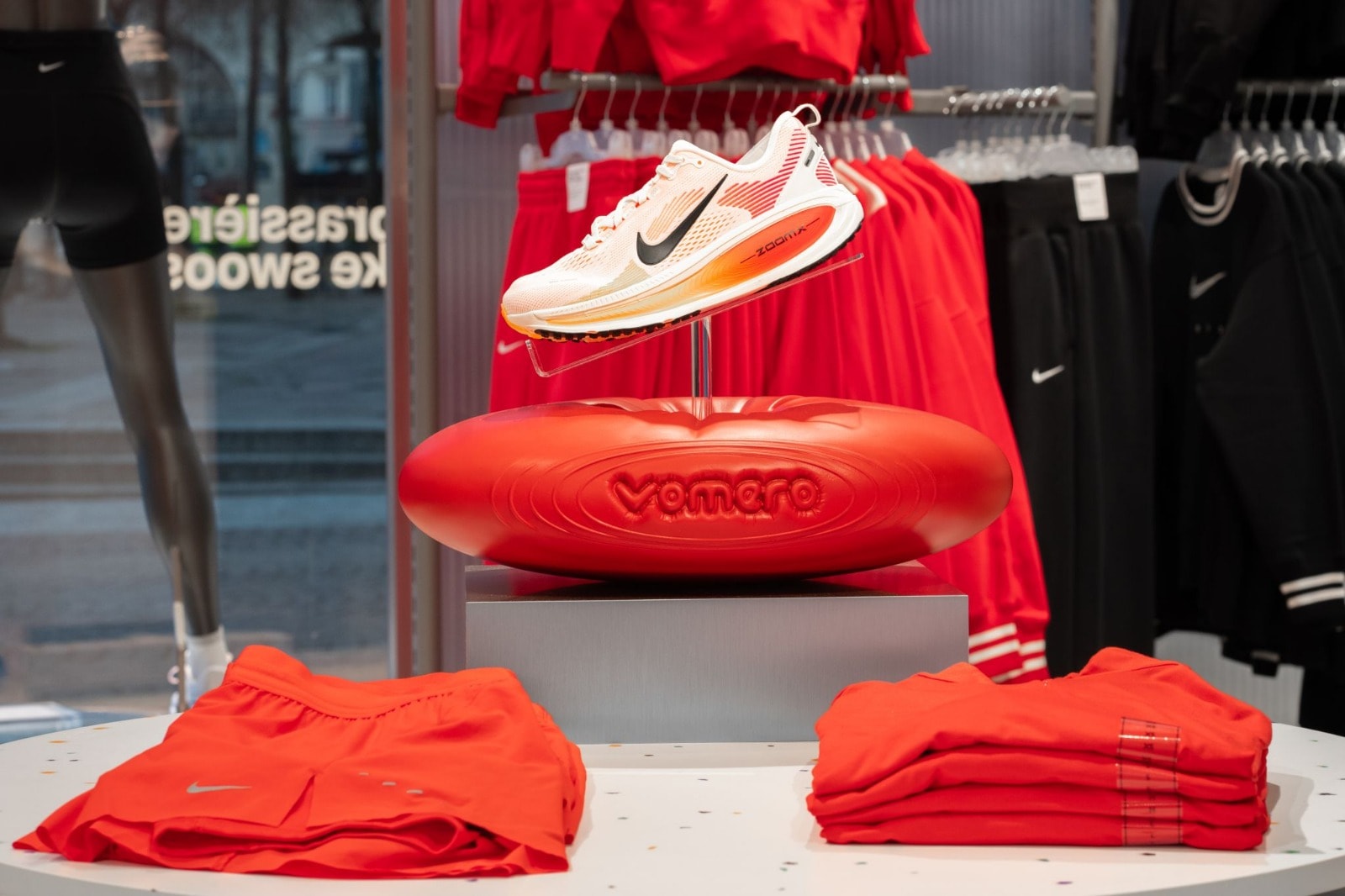 Nike Vomero running shoe showcased on a red branded display pedestal with folded red apparel in a retail store window display.