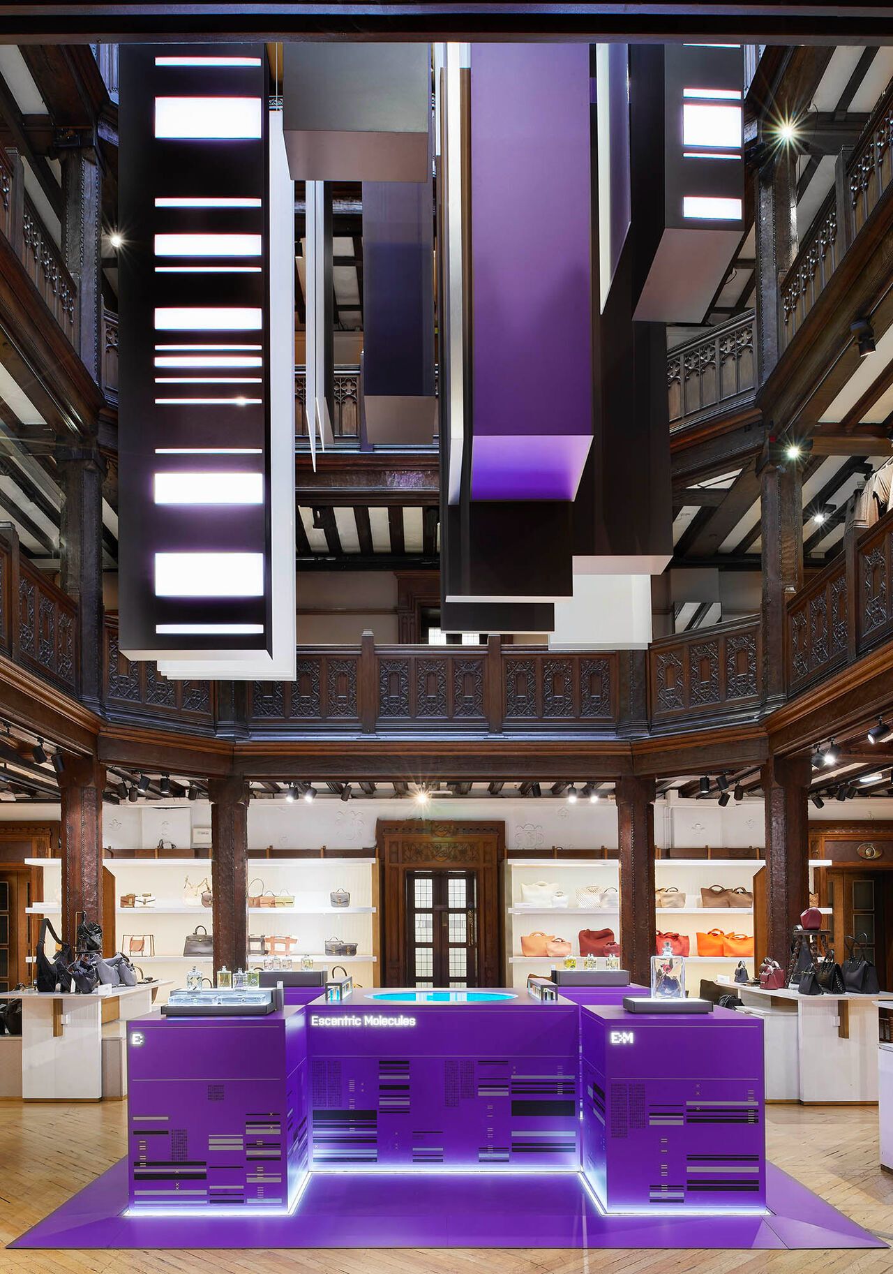 Escentric Molecules purple branded pop-up kiosk and suspended light installation inside an upscale retail interior with handbag displays and illuminated shelving.