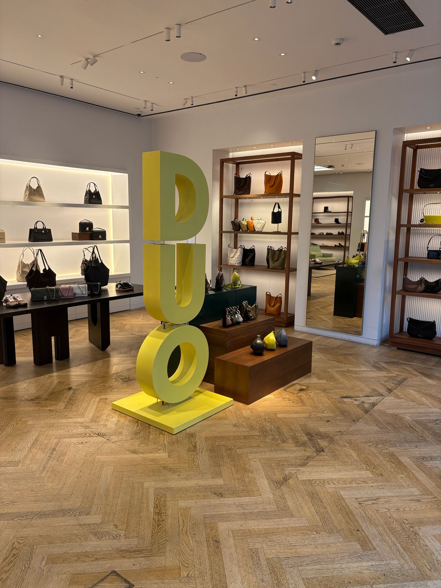 High-end retail store interior showing a freestanding yellow letter display and lit shelving of handbags; ideal example of in-store merchandising and display fixtures.