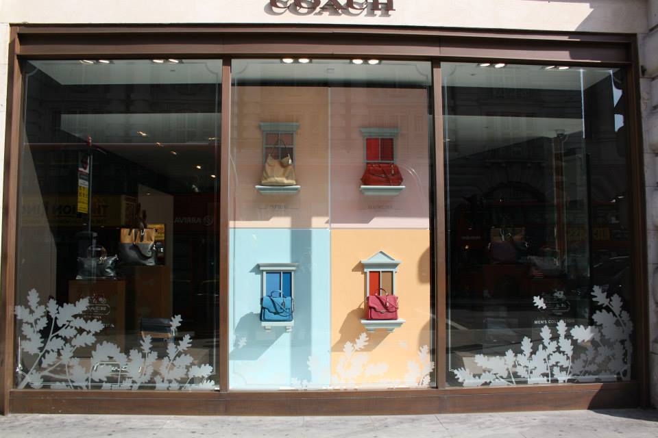Coach retail storefront with branded signage above a window display featuring handbags and decorative vinyl window graphics.