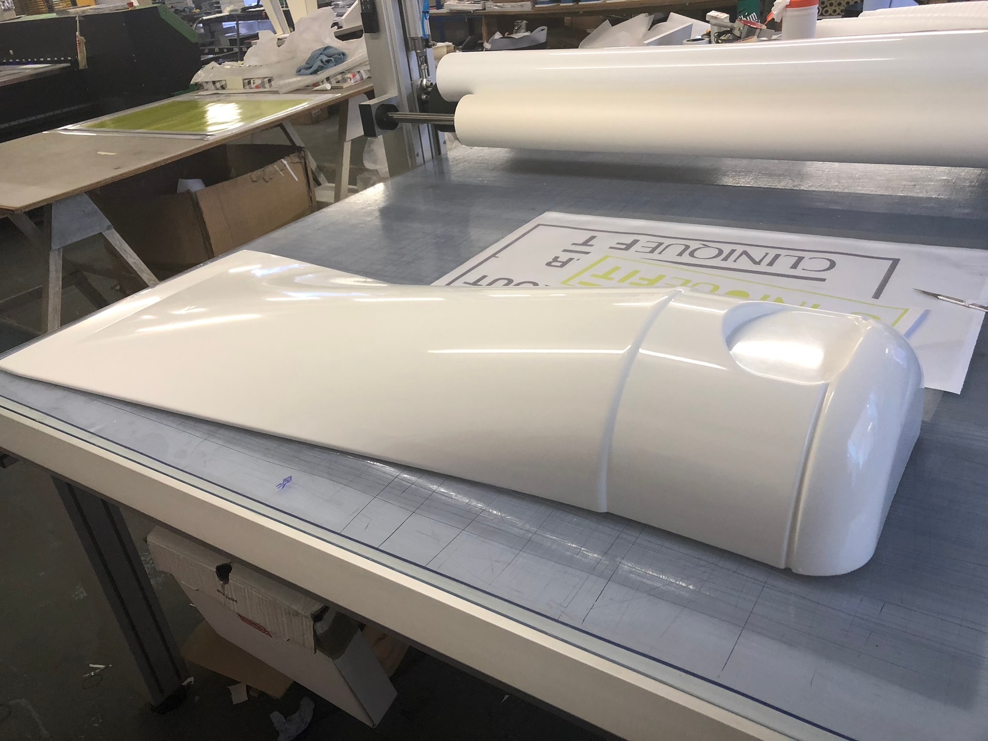 Vacuum-formed white plastic shell on a workshop table with material rolls and printed graphics in the background, showing thermoforming production.